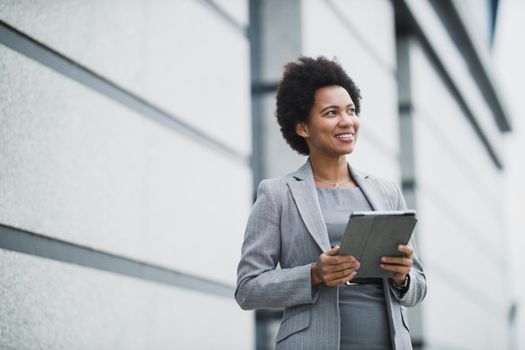 Portrait of a successful black business woman working on a digital tablet during quick break in front a corporate building.