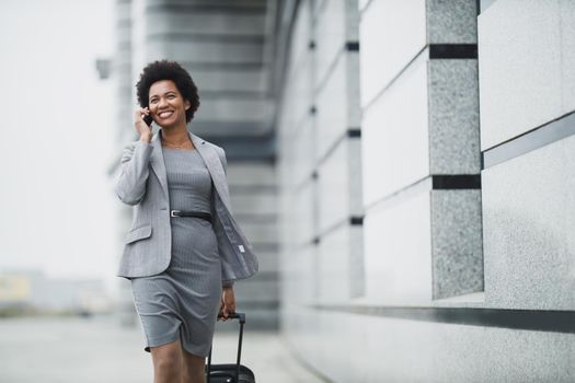 A successful black business woman walking with her luggage and using smart phone outside of the airport.