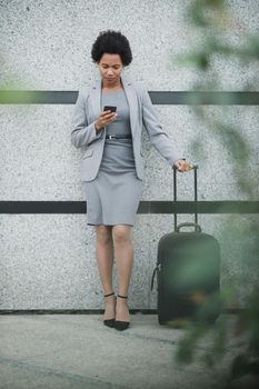A pensive black business woman standing with her luggage and using smart phone outside of the airport.