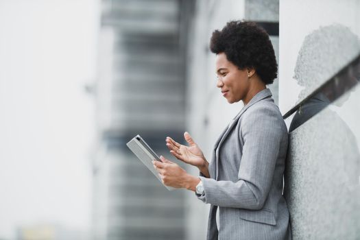 A successful black business woman making video call on a digital tablet during quick break in front a corporate building.