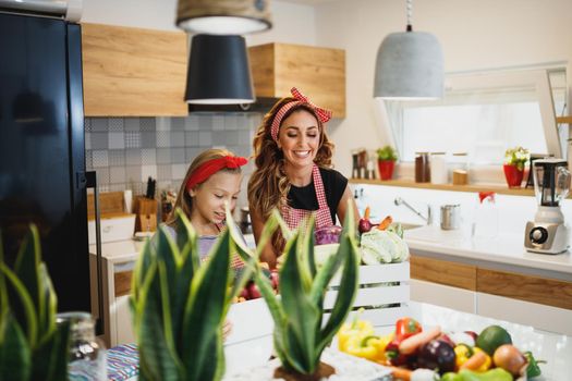 A young cute girl helping her smiling mom preparing fresh vegetables in the theirs kitchen.