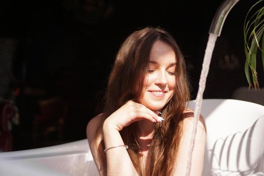 Think positive. Glamorous girl in a bright bathroom with a green plant. A happy woman waits until the bathtub is filled with water