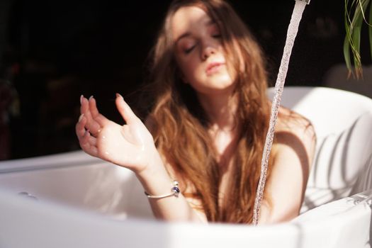 Blurry photo in defocus. Glamorous girl in a bright bathroom with a green plant. A happy woman waits until the bathtub is filled with water