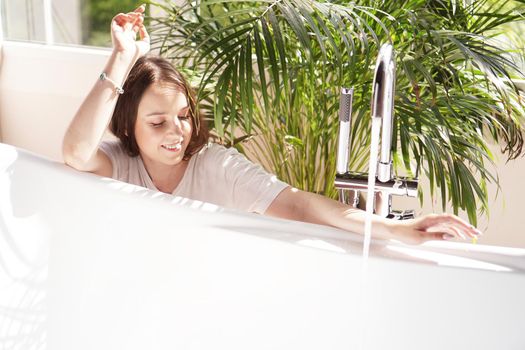 Think positive. Girl in a bright bathroom with a green plant. A happy woman waits until the bathtub is filled with water