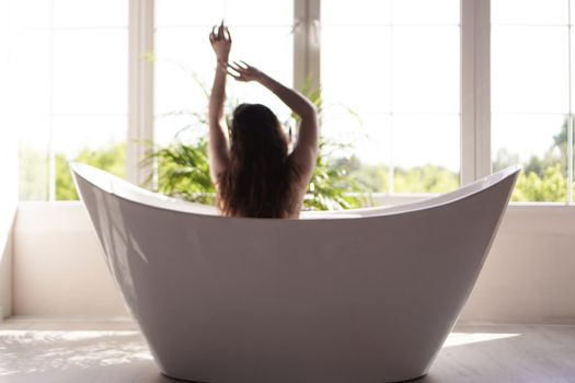 Girl relaxing in bath on light background with big window and sunny light. Blurred photo