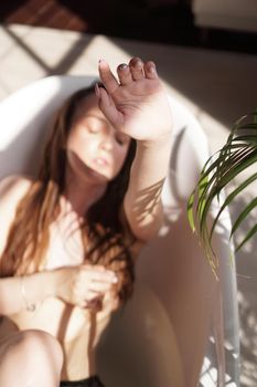 Young attractive woman relaxing in bath - plant in bathroom - sunny day. The model raises the hand - focus on the fingers. Blurred background