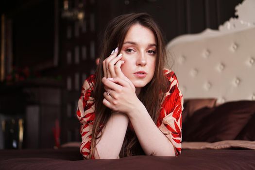 Relaxed woman lying on a brown bed thinking deeply. Woman in retro style room in red dress