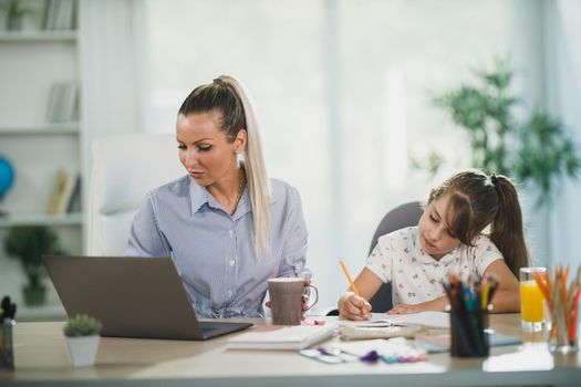 Busy single mother using laptop while her cute daughter learning at home.