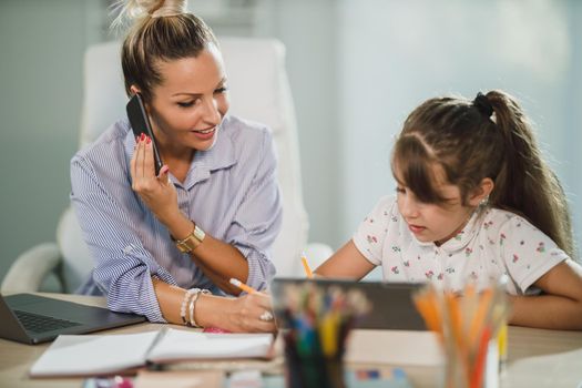 A beautiful busy single mother helping her cute daughter with homework while talking on her smartphone at home.