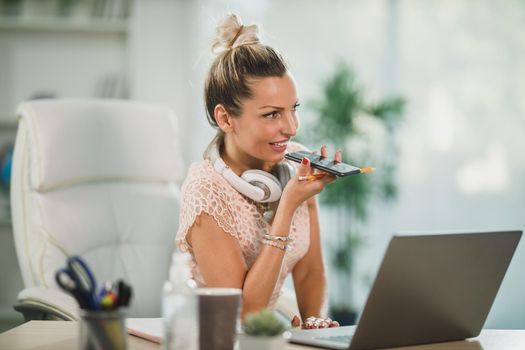 An attractive business woman sitting in her office and sending voice message on her cellphone while working online at her laptop.