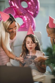 Beautiful young mom with her cute daughters have video call with friends while birthday celebration at home during pandemic isolation.