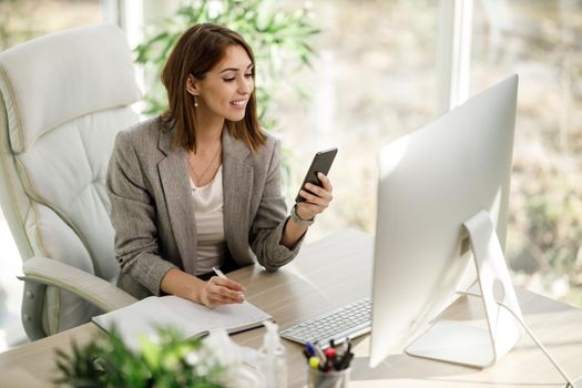 An attractive business woman using smartphone while working at computer in her home office.