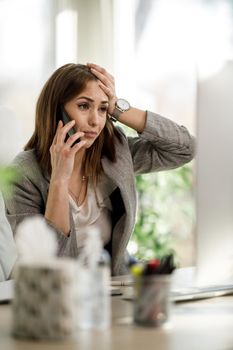 A worried business woman looking stressed out while talking on smartphone and working at computer in her office.