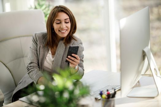 A smiling business woman sitting alone in her home office and using smartphone.