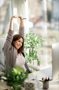 A young business woman looking relaxed with her hands over her head while sitting at her desk and working on computer.
