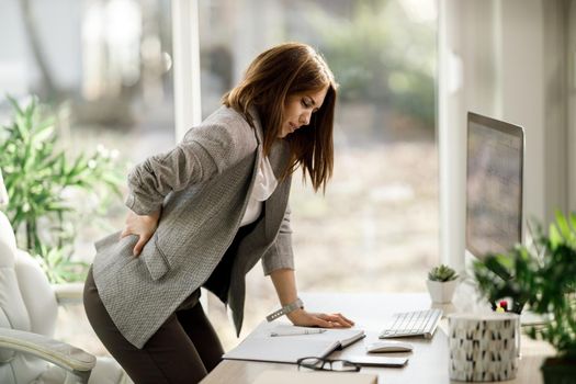 An attractive business woman suffering from back pain while working on computer in a modern office.