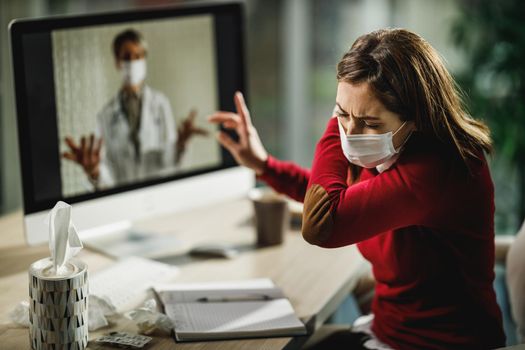 Shot of a young woman coughs at the elbow during a video call with a her doctor on a computer.