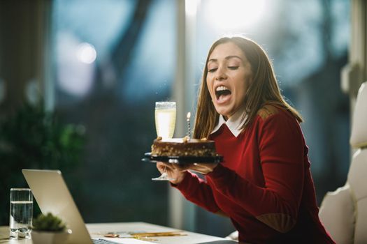 Happy young woman blowing candles on a birthday cake while have a online birthday party with friends.