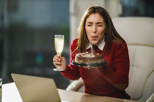 Alone young woman blowing candles on a birthday cake while have a online birthday party with friends.