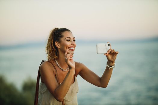 A young cheerful woman making video call on a smartphone while enjoying a summer vacation and exploring the wonderful seaside of Meditteranian.