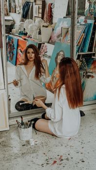 Woman painter sitting on the floor in front of mirror. Art studio interior. Drawing supplies, oil paints, artist brushes