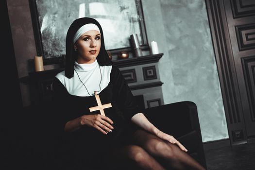 Sexy nun prays indoor . Beautiful young holy sister. Young beautiful nun with a cross in a robe in a black interior.