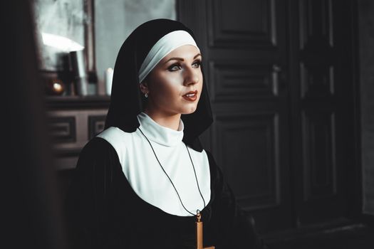 Sexy nun prays indoor . Beautiful young holy sister. Young beautiful nun with a cross in a robe in a black interior.