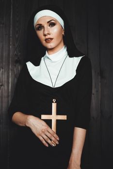 Sexy nun prays indoor . Beautiful young holy sister. Young beautiful nun with a cross in a robe in a black interior.