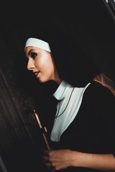 Sexy nun prays indoor . Beautiful young holy sister. Young beautiful nun with a cross in a robe in a black interior.