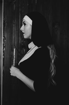 Sexy nun prays indoor . Beautiful young holy sister. Young beautiful nun with a cross in a robe in a black interior. Black and white