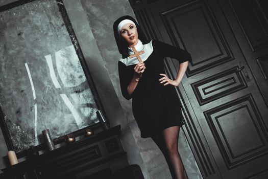 Sexy nun prays indoor . Beautiful young holy sister. Young beautiful nun with a cross in a robe in a black interior.