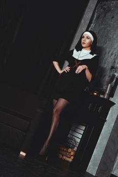 Sexy nun prays indoor . Beautiful young holy sister. Young beautiful nun with a cross in a robe in a black interior.