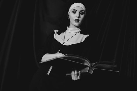 Beautiful young nun in religion black suit holds Bible and posing on camera with big book on a black background. Religion concept. Black and white