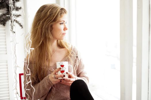 Beautiful young woman drinking coffee and looking through window while sitting at windowsill at home