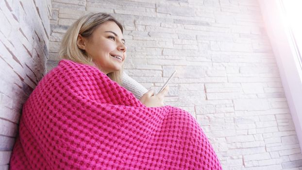 Pretty girl sitting on the window sill with smartphone in hands. She has long blonde hair, smile and looking at window. Pink plaid