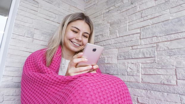 Pretty girl sitting on the window sill with smartphone in hands. She has long blonde hair, smile and looking at her phone. Pink plaid