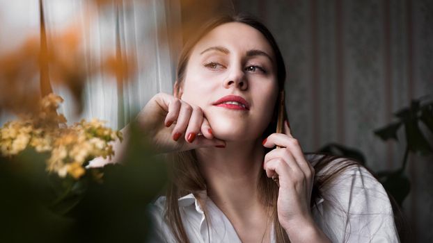 Pretty woman with red lips talking on phone near window at home in morning sunlight