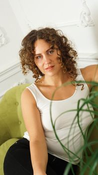 Relaxed lifestyle. Happy young woman with curly hair in white t-shirt spending time at home. Green sofa and white walls in room - vertical photo