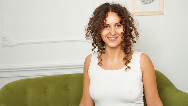 Relaxed lifestyle. Happy young woman with curly hair in white t-shirt spending time at home. Green sofa and white walls in room