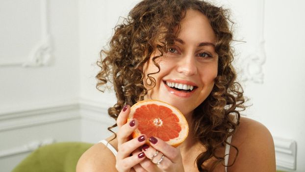 Portrait of pretty middle-aged woman with curly hair with grapefruit at home - light room. The concept of happiness, beauty and health