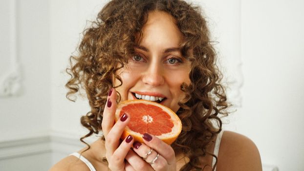 Portrait of pretty middle-aged woman with curly hair with grapefruit at home - light room. The concept of happiness, beauty and health