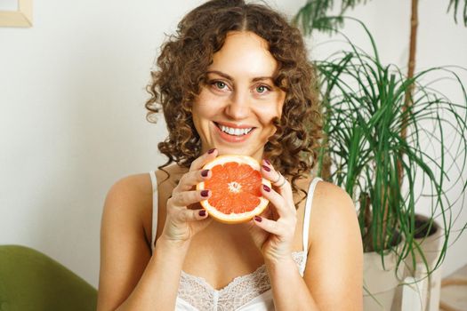 Portrait of pretty middle-aged woman with curly hair with grapefruit at home - light room. The concept of happiness, beauty and health