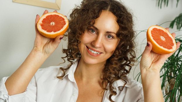 Portrait of pretty middle-aged woman with curly hair with grapefruit at home - light room. The concept of happiness, beauty and health