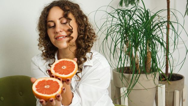 Portrait of pretty middle-aged woman with curly hair with grapefruit at home - light room. The concept of happiness, beauty and health