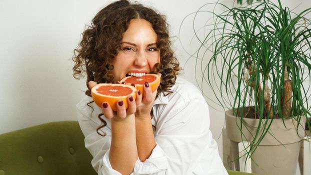 Beautiful woman bites grapefruit. Middle-aged woman with curly hair at home - light room. The concept of happiness, beauty and health