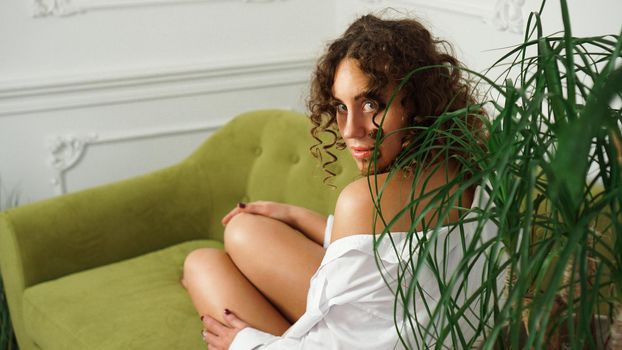 Woman sitting on green sofa in living room. Beautiful long legs. Beautiful woman with curly hair in white lingerie at home - happy morning