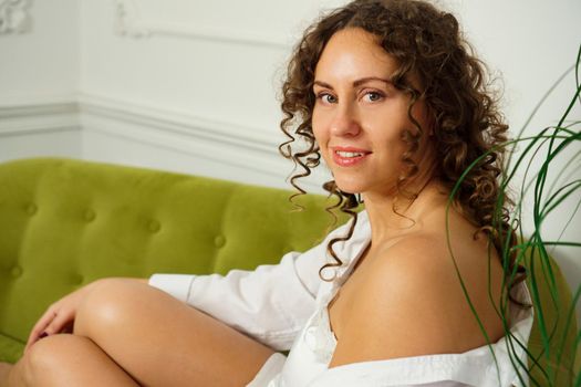 Relaxed lifestyle. Happy young woman with curly hair in white shirt spending time at home. Green sofa and white walls in room