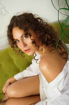 Relaxed lifestyle. Happy young woman with curly hair in white shirt spending time at home. Green sofa and white walls in room