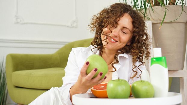 Beauty cosmetics concept. Beautiful young woman with curly hair and healthy skin in room with a green sofa. Apples and Organic Care on a wooden table