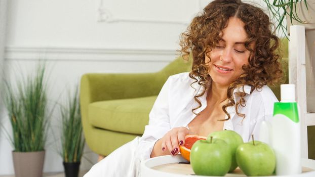 Beauty cosmetics concept. Beautiful young woman with curly hair and healthy skin in room with a green sofa. Apples and Organic Care on a wooden table